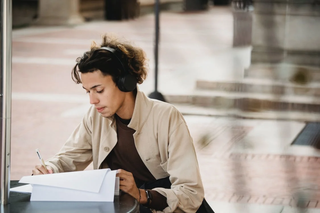 How Technology Helps Writers Stay Organized And Productive 1 Ethnic female student in earbuds writing on copybook near laptop